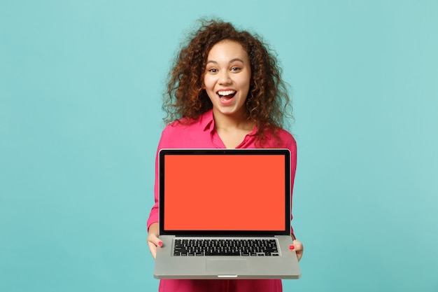 Opgewonden Afrikaans meisje in casual kleding houdt laptop pc-computer met leeg leeg scherm geïsoleerd op blauwe turkooizen achtergrond in studio. Mensen oprechte emoties, lifestyle concept. Bespotten kopie ruimte.