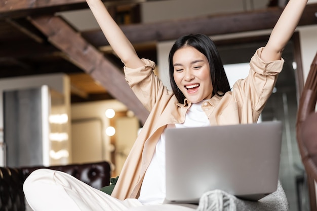 Opgewonden Aziatisch meisje wint online op laptop en zegt ja hoera en viert thuis zitten