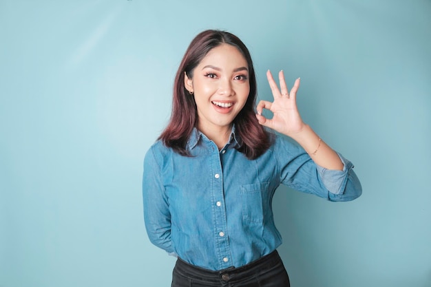 Opgewonden Aziatische vrouw die een blauw overhemd draagt dat een OK handgebaar geeft dat door een blauwe achtergrond wordt geïsoleerd