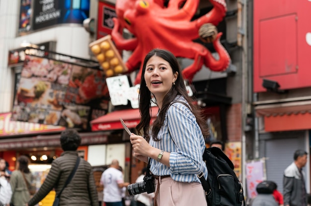 opgewonden Aziatische vrouw die rondkijkt met een kaart in de hand terwijl hij geniet van een actieve sfeer in het winkelgebied in Shinsaibashisuji en Dotonboshiri Osaka Japan met een octopusbord erachter