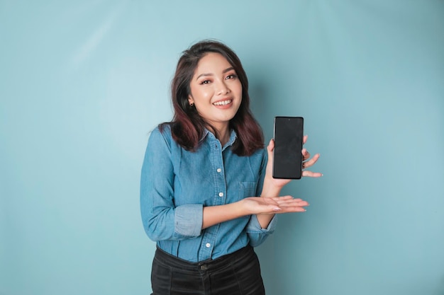 Opgewonden Aziatische vrouw met een blauw shirt wijzend op de kopieerruimte op haar smartphone geïsoleerd door blauwe achtergrond