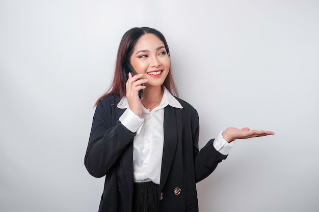 Opgewonden Aziatische zakenvrouw in zwart pak wijzend naar de kopieerruimte naast haar terwijl ze aan het praten is op haar telefoon geïsoleerd door een witte achtergrond