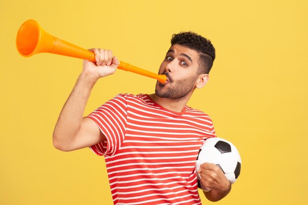 Opgewonden bebaarde man in rood gestreept t-shirt blazen in hoorn met zwart-wit voetbal, vieren overwinning van favoriete voetbalteam. Indoor studio-opname geïsoleerd op gele achtergrond