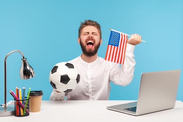 Opgewonden bebaarde man voetbal fanat oprecht vreugde ondersteunende favoriete Amerikaanse team houden voetbal en usa vlag kijken wedstrijd op werkplek Indoor studio shot geïsoleerd op blauwe achtergrond