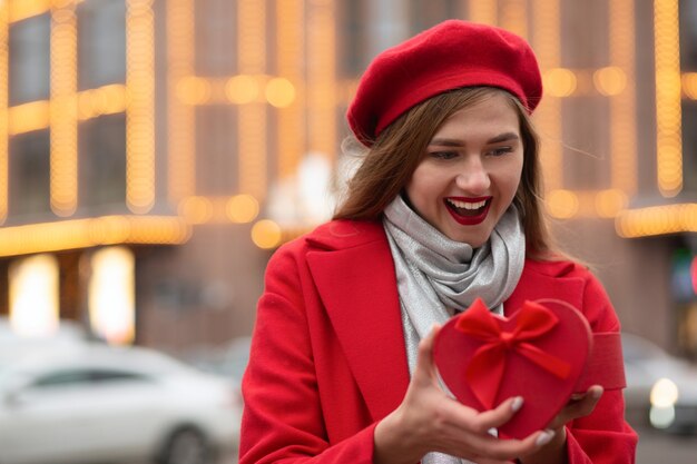 Opgewonden blonde vrouw draagt een rode baret en een jas die een hartvormige geschenkdoos opent op de achtergrond van bokehlichten. ruimte voor tekst