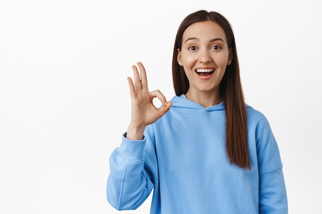 Opgewonden brunette vrouw toont oké, nul OK-teken en glimlacht geamuseerd, ben het met je eens, vind het leuk en keur goed, prijs geweldig ding, staande tegen een witte muur