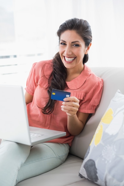 Opgewonden brunette zittend op haar bank met behulp van laptop om online te winkelen