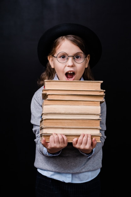 Opgewonden elementair schoolmeisje in hoed en bril die stapel boeken tegen zwarte ruimte afzonderlijk dragen