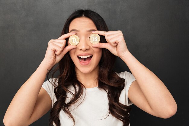 Opgewonden emotionele Aziatische jonge vrouw die twee gouden bitcoins voor ogen.