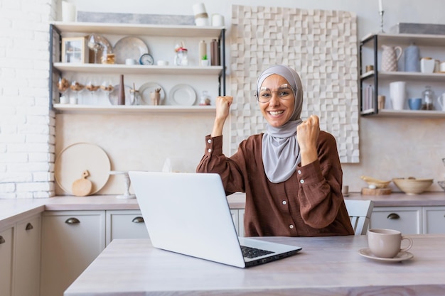 Opgewonden en gelukkige moslim jonge vrouw in hijab om thuis te zitten met behulp van laptop en het vieren van succes