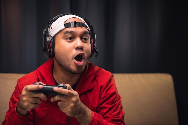 Opgewonden en geschokt gezicht van Aziatische gamer met hoofdtelefoon met headset die videogame online speelt zittend op de bank in de woonkamer Indiase professionele gamer die op sociale speelgame streamt, erg leuk