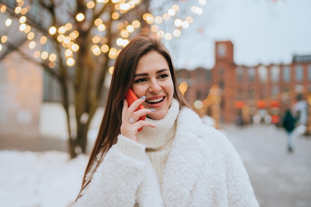 Opgewonden Europees meisje in witte bontjas praat via de telefoon staat buiten tegen wazige bloemenslingers