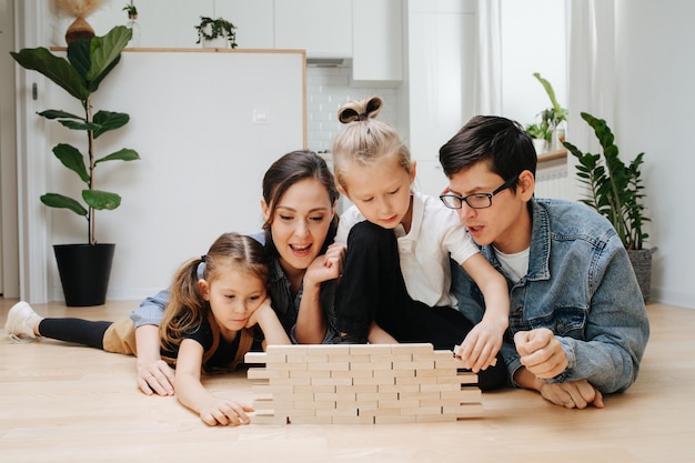 Opgewonden familie die jenga op de vloer speelt en tegels uit een houten blokmuur trekt Ouders en kinderen die op de vloer liggen