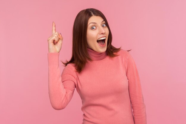 Opgewonden geïnspireerde vrouw met bruin haar wijzende vinger omhoog kijkend naar camera met brede glimlach, blij met nieuw geweldig idee, oplossing vinden. Indoor studio-opname geïsoleerd op roze background