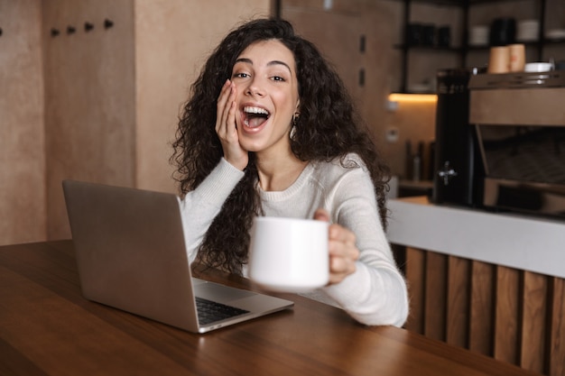 Opgewonden gelukkige vrouw die laptopcomputer gebruikt terwijl ze aan de cafétafel zit en koffie drinkt