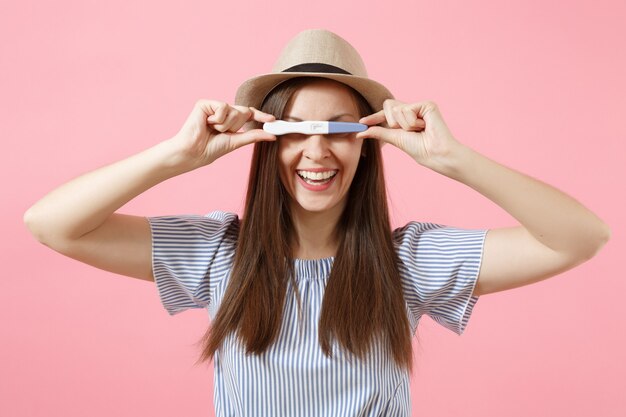 Opgewonden gelukkige vrouw in blauwe jurk, hoed in de hand houden, kijken naar zwangerschapstest geïsoleerd op roze achtergrond. medische gezondheidszorg gynaecologische, zwangerschap vruchtbaarheid moederschap mensen concept. ruimte kopiëren.