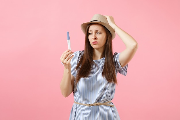 Opgewonden gelukkige vrouw in blauwe jurk, hoed in de hand houden, kijken naar zwangerschapstest geïsoleerd op roze achtergrond. Medische gezondheidszorg gynaecologische, zwangerschap vruchtbaarheid moederschap mensen concept. Ruimte kopiëren.