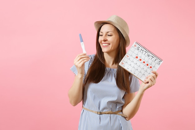 Opgewonden gelukkige vrouw in blauwe jurk, hoed in de hand houden zwangerschapstest, periodenkalender voor het controleren van menstruatiedagen geïsoleerd op roze achtergrond. Medisch, gezondheidszorg, gynaecologisch concept. Ruimte kopiëren.