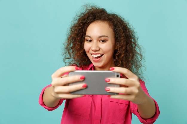 Foto opgewonden grappig afrikaans meisje in roze casual kleding doen selfie schot op mobiele telefoon geïsoleerd op blauwe turquoise muur achtergrond in studio. mensen oprechte emoties levensstijl concept. bespotten kopie ruimte.