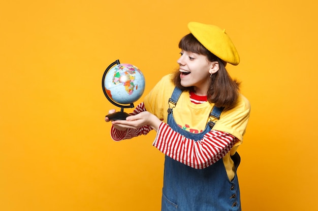 Opgewonden grappig meisje tiener in Franse baret en denim zomerjurk met Earth wereldbol geïsoleerd op gele muur achtergrond in studio. Mensen oprechte emoties, lifestyle concept. Bespotten kopie ruimte.