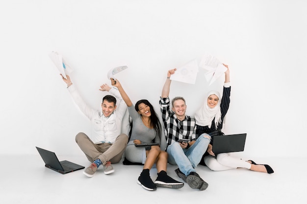 Opgewonden groep van vier multi-etnische vrienden die handen opheffen, die samen op de vloer op witte achtergrond zitten