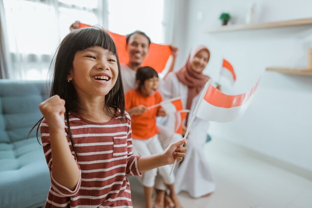 Opgewonden indonesische aanhanger van het kleine meisje tijdens het kijken naar sportwedstrijd op televisie thuis met familie op de achtergrond