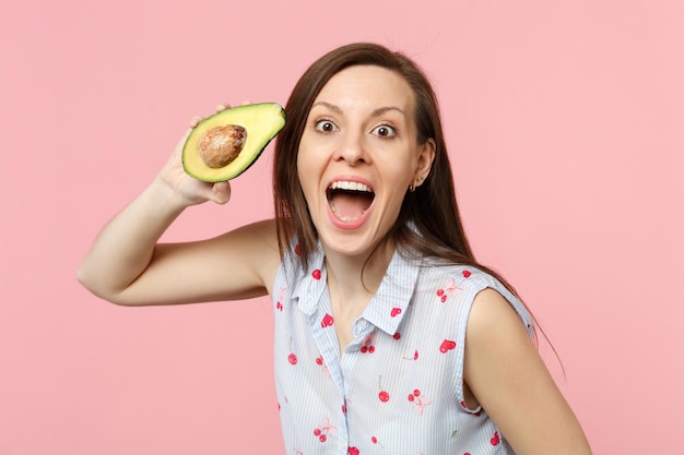 Opgewonden jong meisje in zomerkleren die mond open houden, houd vers rijp avocadofruit geïsoleerd op roze pastelachtergrond in studio. Mensen levendige levensstijl ontspannen vakantie concept. Bespotten kopie ruimte.