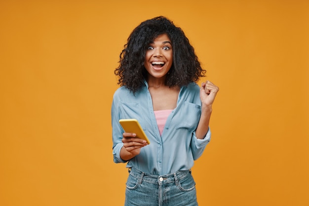 Opgewonden jonge Afrikaanse vrouw in casual kleding met behulp van slimme telefoon en glimlachen