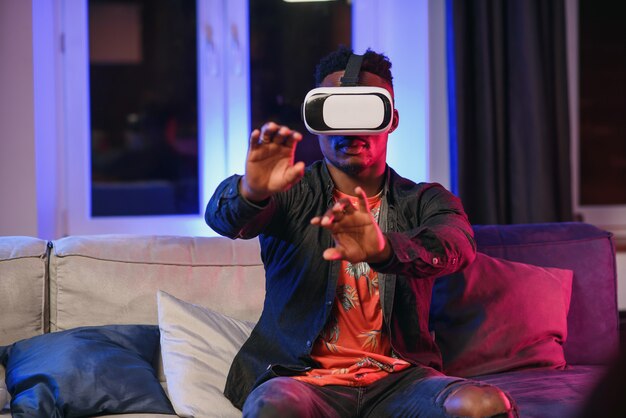 Opgewonden jonge afro-amerikaanse man hand vooruit uit te breiden, met behulp van virtual reality headset zittend op bed, kopie ruimte