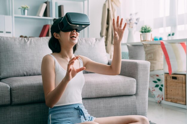 Opgewonden jonge aziatische japanse vrouw in casual kleding die thuis ontspant in een VR-bril terwijl ze op de vloer van de woonkamer zit en de lucht met de hand aanraakt. charmant meisje dat een virtual reality-bril draagt.