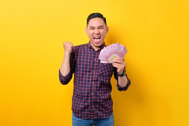 Opgewonden jonge Aziatische man die geld bankbiljetten vasthoudt en vuist viert financieel succes geïsoleerd op gele achtergrond Winst en rijkdom concept