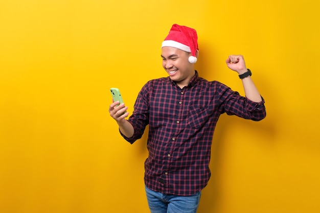 Foto opgewonden jonge aziatische man in kerstmuts met behulp van smartphone en winnaar gebaar doen over gele studio achtergrond gelukkig nieuwjaar 2023 viering vrolijk vakantie concept