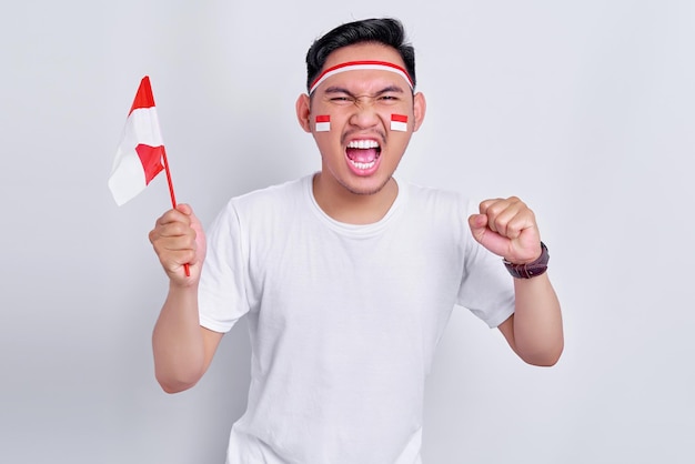 Opgewonden jonge aziatische man met indonesische vlag en balde beide vuisten om de onafhankelijkheidsdag van indonesië te vieren op 17 augustus geïsoleerd op een witte achtergrond