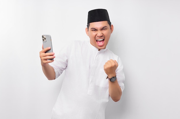 Opgewonden jonge Aziatische moslim man met behulp van mobiele telefoon en het verhogen van de vuist het vieren van geluk geïsoleerd op witte achtergrond Mensen religieus Islam lifestyle concept viering Ramadan en ied Mubarak