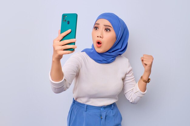 Opgewonden jonge Aziatische moslimvrouw die smartphone gebruikt en winnaargebaar doet geïsoleerd op witte achtergrond