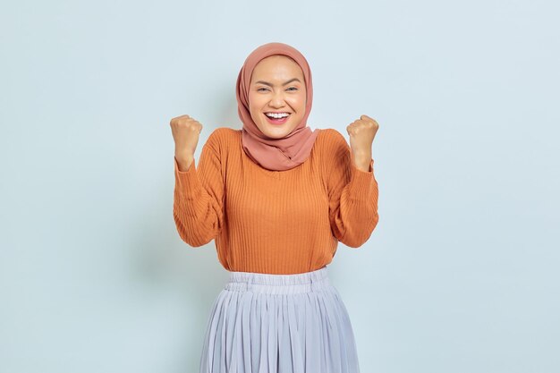 Opgewonden jonge Aziatische moslimvrouw in bruine trui staande doen winnend gebaar vieren vuist ja zeggen geïsoleerd op witte achtergrond moslim lifestyle concept