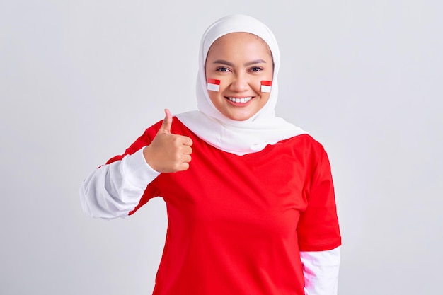 Opgewonden jonge Aziatische moslimvrouw in rood wit tshirt vieren Indonesische onafhankelijkheidsdag op 17 augustus en tonen duim omhoog gebaar met vingers geïsoleerd op witte achtergrond