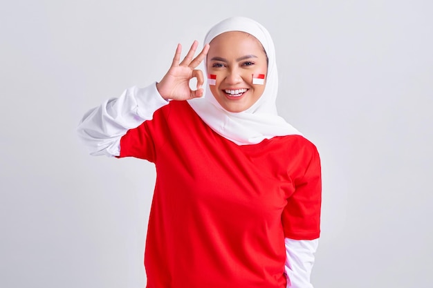 Opgewonden jonge Aziatische moslimvrouw in rood wit tshirt vieren Indonesische onafhankelijkheidsdag op 17 augustus en tonen oke gebaar geïsoleerd op witte achtergrond