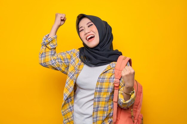 Opgewonden jonge Aziatische moslimvrouw student in casual kleding en rugzak doen winnaar gebaar geïsoleerd op gele achtergrond Onderwijs school universiteit college concept