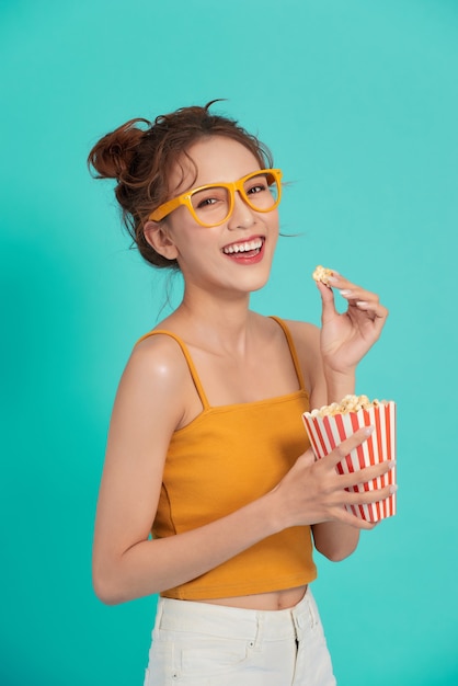 Opgewonden jonge Aziatische vrouw die popcorn eet die op blauw wordt geïsoleerd.