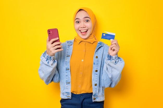 Opgewonden jonge Aziatische vrouw in jeans jasje met mobiele telefoon en creditcard online winkelen geïsoleerd over gele achtergrond