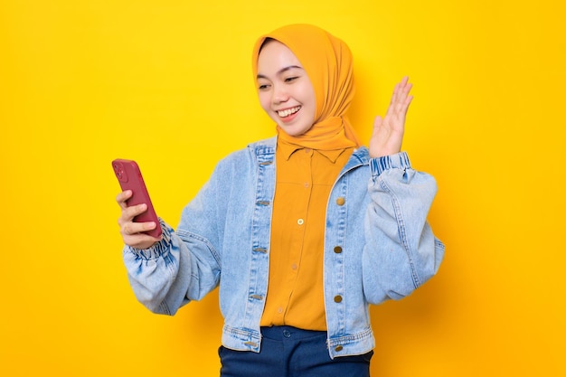 Opgewonden jonge Aziatische vrouw in spijkerjasje die naar het scherm van de mobiele telefoon kijkt, voelt zich vrolijk bij het lezen van goed nieuws geïsoleerd over gele achtergrond