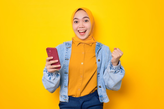 Opgewonden jonge Aziatische vrouw in spijkerjasje met behulp van mobiele telefoon die succes viert en goed nieuws krijgt geïsoleerd over gele achtergrond