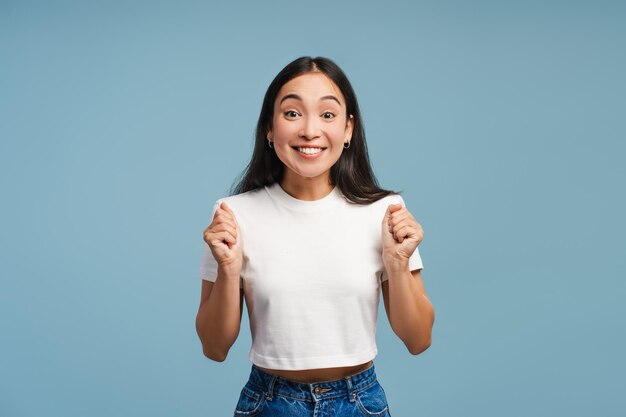 Opgewonden jonge Aziatische vrouw viert succes met vuisten omhoog op blauwe achtergrond