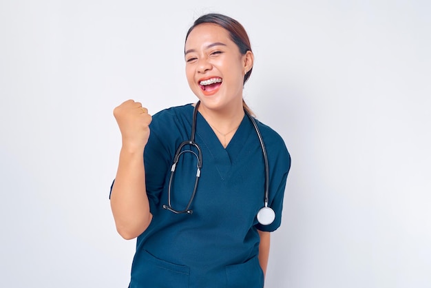 Foto opgewonden jonge aziatische vrouwelijke professionele verpleegster die een blauw uniform draagt en zelfverzekerd staat terwijl ze ja zegt en de overwinning viert geïsoleerd op witte achtergrond gezondheidszorg geneeskunde concept