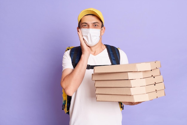 Opgewonden jonge blanke bezorger in beschermend masker met pizzadozen geïsoleerd op paarse achtergrond kijkend naar camera met verbazing houdt hand op wang kijkend naar camera