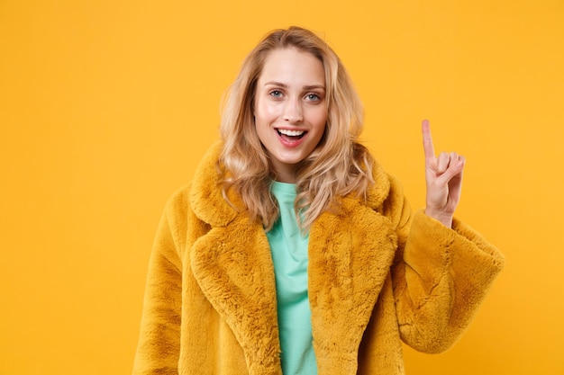 Opgewonden jonge blonde vrouw meisje in gele bontjas poseren geïsoleerd op oranje muur achtergrond studio portret. Mensen levensstijl concept. Bespotten kopie ruimte. Wijsvinger omhoog houden met geweldig nieuw idee.