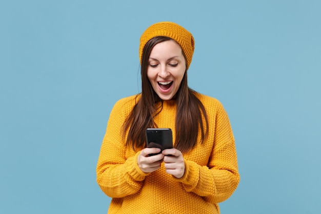 Opgewonden jonge brunette vrouw meisje in gele trui en hoed poseren geïsoleerd op blauwe achtergrond studio portret. Mensen levensstijl concept. Bespotten kopie ruimte. Met behulp van mobiele telefoon, sms-bericht typen.