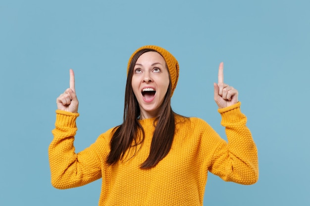 Opgewonden jonge brunette vrouw meisje in gele trui hoed poseren geïsoleerd op blauwe achtergrond studio portret. Mensen oprechte emoties levensstijl concept. Bespotten kopie ruimte. Wijsvingers omhoog wijzen.