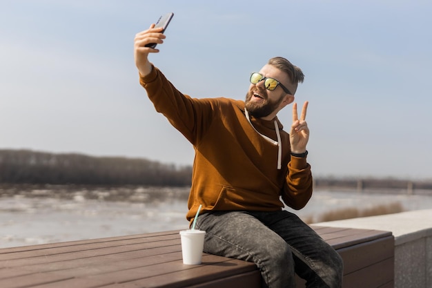 Opgewonden jonge kerel maakt selfie op smartphone, hij is gen z of millennial en draagt casual trendy